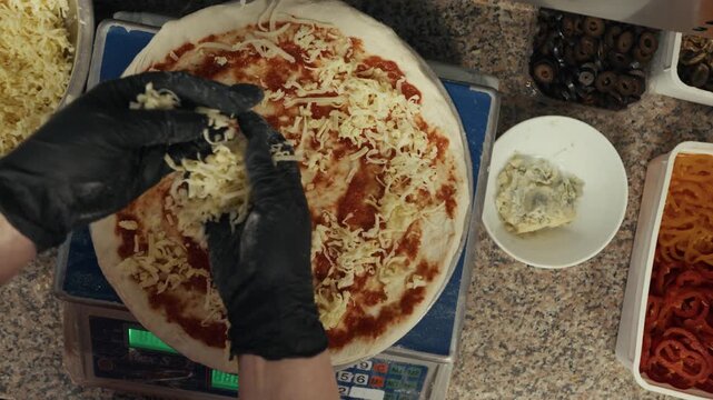 Hands of a chef in black gloves sprinkle cheese onto a pizza base with tomato sauce, surrounded by various toppings in a commercial kitchen setting
