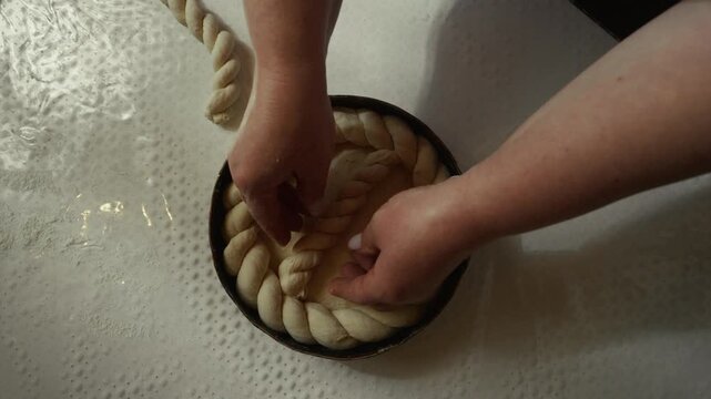 Woman preparing braided bread dough in a round baking pan, hands skillfully arranging the dough on a clean countertop with flour visible in the background