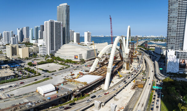 Aerial view of the signature arches of a bridge under construction contrasting against the modern skyline and Biscayne Bay, Miami, Florida, USA.