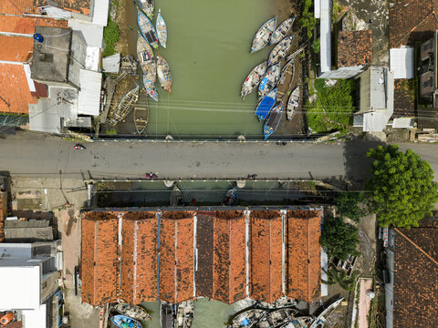 Aerial view of boats resting in the river near the market and bridge, contrasting with the vibrant orange roofs of the buildings, Jepara, Central Java, Indonesia.