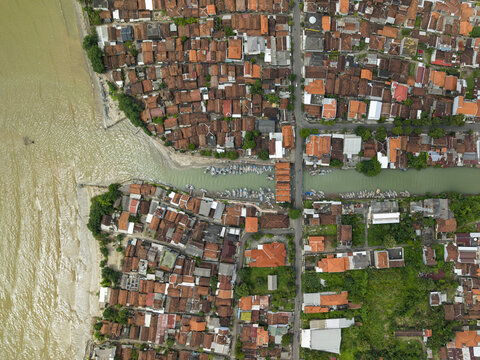 Aerial view of terracotta rooftops meet the murky waters at the Jepara Floating Fish Market, contrasting with the boats docked along the river, Jepara, Central Java, Indonesia.