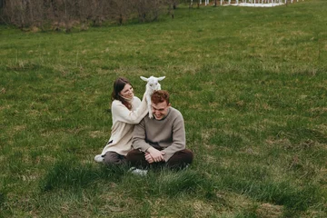 Fototapete Tierärzte Cheerful couple outdoors, sitting on grass, playful goat kid beside them, relaxed mood, casual clothing, soft light, natural colors, rural meadow backdrop  © SHOTPRIME STUDIO