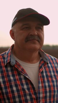 Middle aged man in plaid shirt and cap standing outdoors at sunset, thoughtful expression on face as he looks into distance in warm natural countryside light