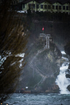 View of a powerful waterfall cascading down the rocks, mist swirling around the Swiss flag atop a stone perch, Neuhausen am Rheinfall, Switzerland.