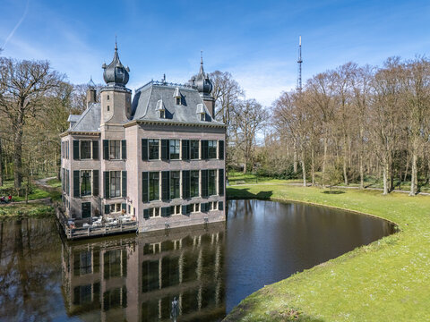 Aerial view of the historic Landgoed Oud Poelgeest castle reflecting in the still waters, framed by spring trees under a vast blue sky, Oegstgeest, Zuid-Holland, Netherlands.
