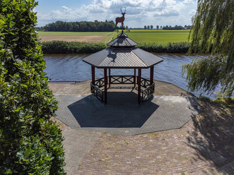 Aerial view of a gazebo topped with a deer statue, standing proudly against a backdrop of green fields and a tranquil canal, Tollebeek, Flevoland, Netherlands.