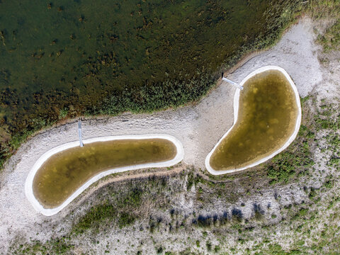 Aerial view of kidney shaped ponds with concrete borders, reflecting sunlight in their shallow waters, surrounded by sandy banks and green vegetation, Almere, Flevoland, Netherlands.