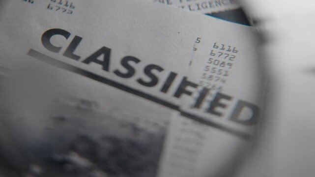 Classified document close up on official government report with magnifying glass. Top secret sensitive information or intelligence revealed and exposed. Office desk with folders and confidential files