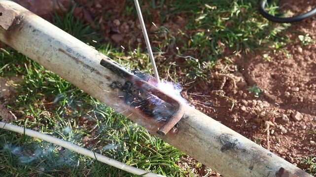 A welder is performing arc welding on an iron pipe in an open area.