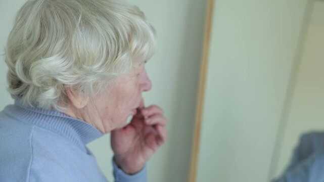 Elderly woman looking at herself in mirror and touching face, examining wrinkles. Authentic lifestyle moment reflecting aging, self perception and natural beauty.
