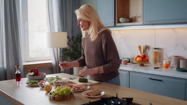 Aged woman puts prosciutto and leaves onto crispbread in kitchen