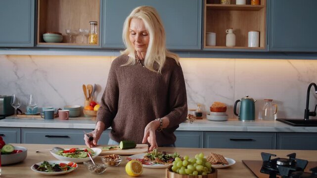Happy old woman adds cucumber slices to salad at wooden counter