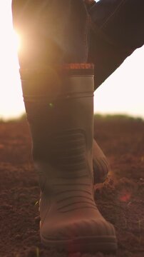 Close-up view of a person wearing rugged rubber boots walking across freshly tilled soil in a rural agricultural field at sunrise, symbolizing farming and hard work outdoors
