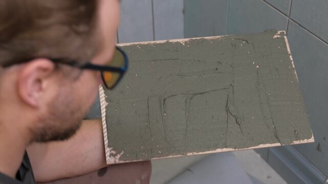 Tile adhesive spreading. Tiling mortar application. Worker smoothing adhesive. Workman smoothing grout on ceramic tiles. Bathroom renovation. Man using notched trowel, cement spread across tile back