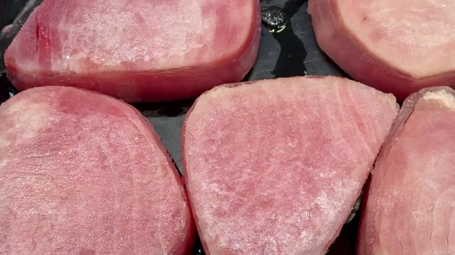 Pink tuna steaks on black tray, supermarket shelf closeup, chilled display of raw fresh fillets arranged in neat rows, subtle.