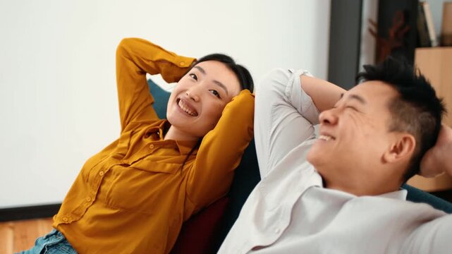 A man with dark hair and a woman with long black hair sit comfortably on a couch in a modern living room. They smile and lean back, showing enjoyment of their time together.