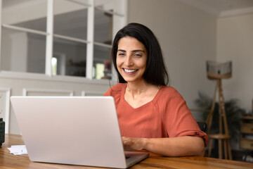 Fototapeta premium Cheerful Brazilian woman sits at desk, smiling while working on laptop in modern homeoffice, make purchase through website, ordering goods, using electronic commerce services at home. Tech, internet