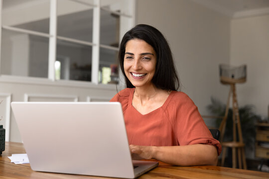 Cheerful Brazilian woman sits at desk, smiling while working on laptop in modern homeoffice, make purchase through website, ordering goods, using electronic commerce services at home. Tech, internet