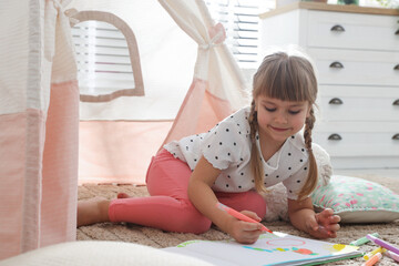 Little girl drawing in sketchbook near toy wigwam at home © New Africa