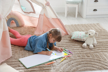 Little girl drawing in sketchbook near toy wigwam at home © New Africa