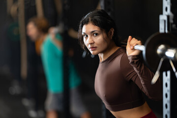 Fototapeta premium Determined Latina girl working out with barbell in well-equipped gym during training session