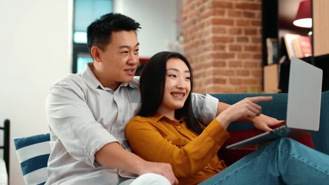 A man with light skin sits behind a woman with light skin on a couch, both smiling. They are looking at a laptop on their laps in a bright living room.