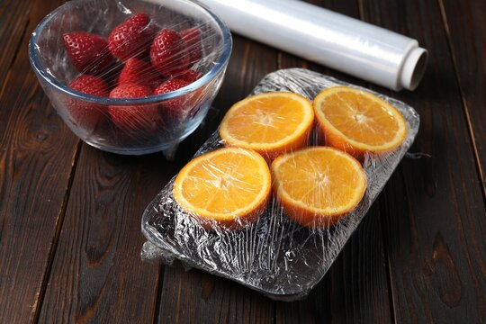 Fresh fruits wrapped in cling film and roll of plastic wrap on wooden table, closeup