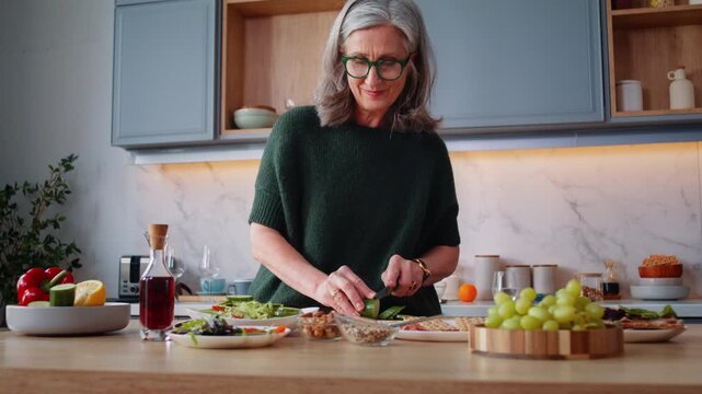 Old woman wearing spectacles cuts cucumber as cooks at counter