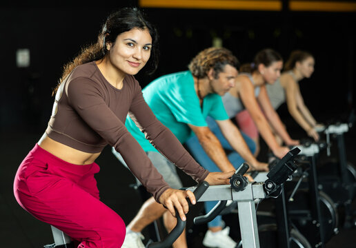Positive Latina girl training with spin bike in fitness hall during full-body workout