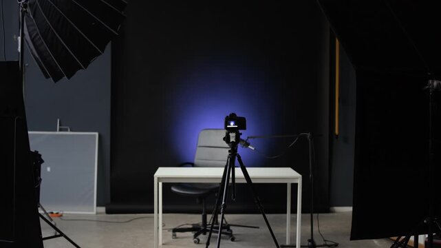 Spacious modern studio with professional equipment. Table and chair placed in center of room with jet black backdrop in background, professional camera and lamps in foreground, cope space