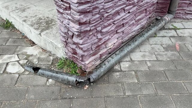 Cracked pipe by brick corner leaking onto pavement, corroded metal downspout detached from wall, small hole releasing water, moss.