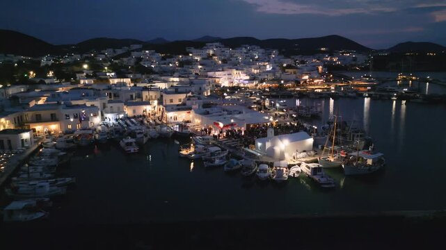 Evening aerial drone view of Naousa village on Paros island, Greece, featuring illuminated streets, coastal harbor and festive atmosphere during a local celebration. Ideal for travel, culture