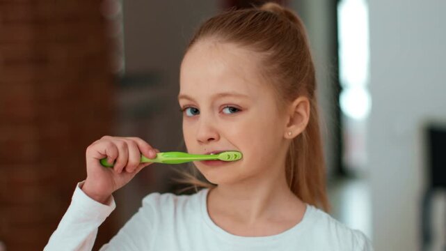 A young girl with light skin and long hair brushes her teeth with a green toothbrush. She smiles while standing in a modern home, demonstrating good dental hygiene habits.