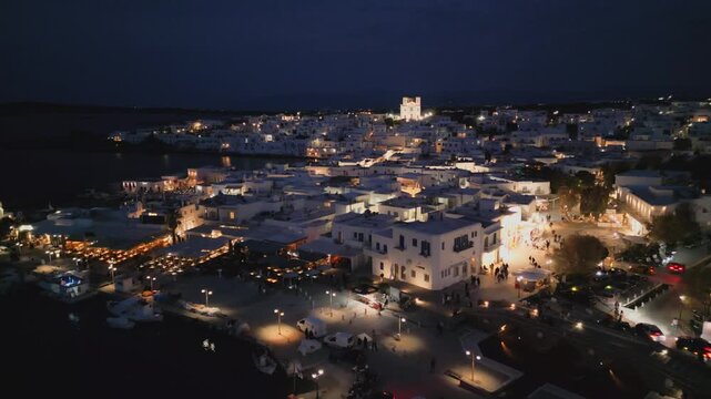 Evening aerial drone view of Naousa village on Paros island, Greece, featuring illuminated streets, coastal harbor and festive atmosphere during a local celebration. Ideal for travel, culture