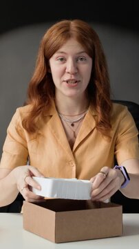 Vertical shot of red haired young woman sitting at table, unpacking brand new wireless headphones while recoding new video for blog