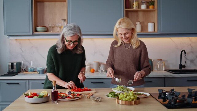 Cheerful senior women talk cooking salad together in kitchen