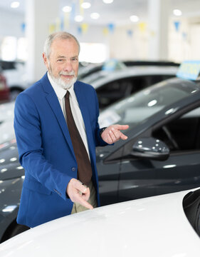 Elderly car salesman stands in car dealership and offers various cars to potential buyers