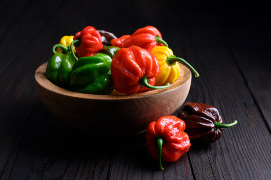Bowl filled with habanero peppers on dark wooden table with one outside. Fresh chili assortment red green yellow varieties rustic kitchen scene