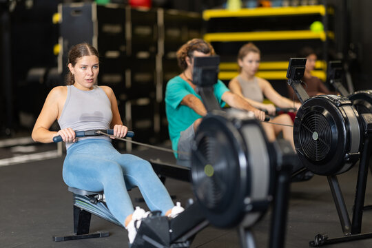 Young woman with sports leggings and a top does a cardio workout on a rowing machine. Crossfit group training participants perform a circuit training session for endurance.