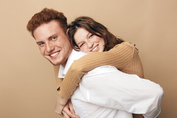 Fototapeta premium Smiling young couple in casual clothes embraces happily against beige background. The woman hugs the man from behind, both showing joyful expressions. Concept of love, friendship, and togetherness.