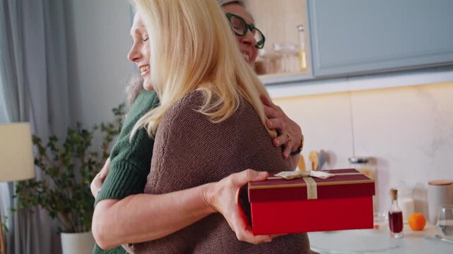 Senior woman gives red giftbox to surprised friend in kitchen