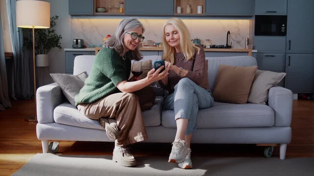 Smiling aged women scroll social media on smartphone on sofa