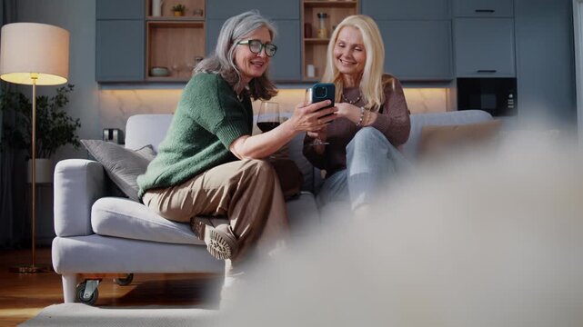 Senior female friends watch smartphone together smiling on sofa
