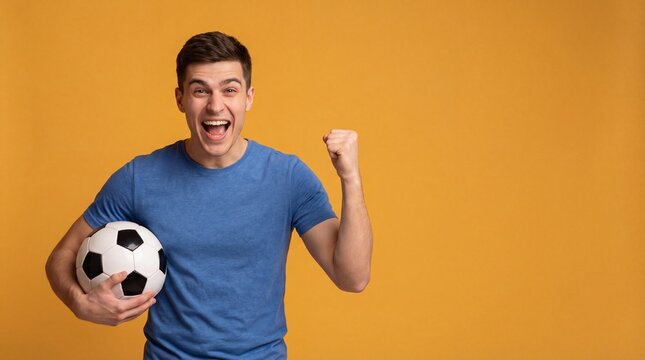 Excited young man holding a soccer ball and celebrating victory against a vibrant orange backdrop