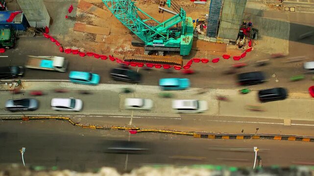 Timelapse of Vehicles move around the busy construction zone at Matraman area. Heavy machinery and steel beams dominate the scene. Workers manage equipment amid greenery and urban buildings.