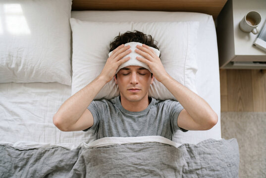 Sick young man lying in bed holding a warm compress to his forehead to relieve a fever