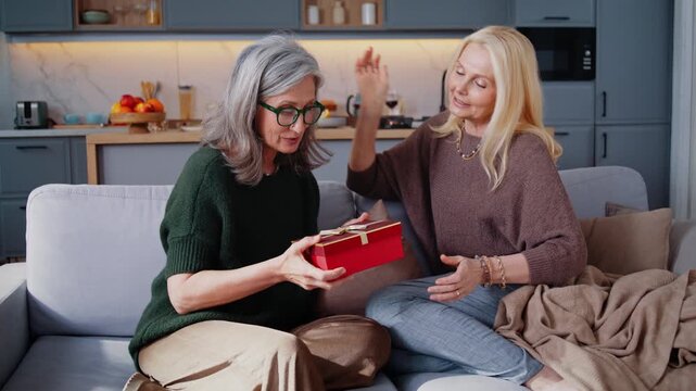 Mature woman receives red gift box from blonde friend on sofa