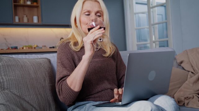 Elderly blonde woman drinks red wine while using laptop on sofa