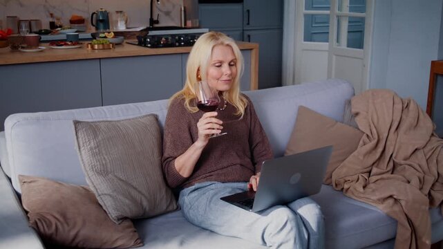 Blonde senior woman drinks wine using laptop on sofa at home