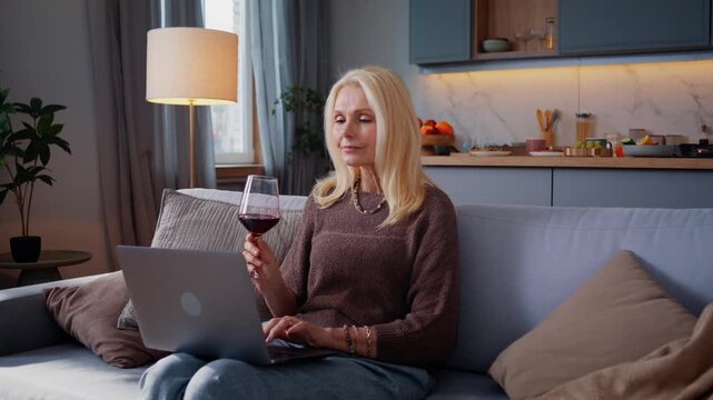 Mature woman sips red wine pausing evening laptop work on sofa
